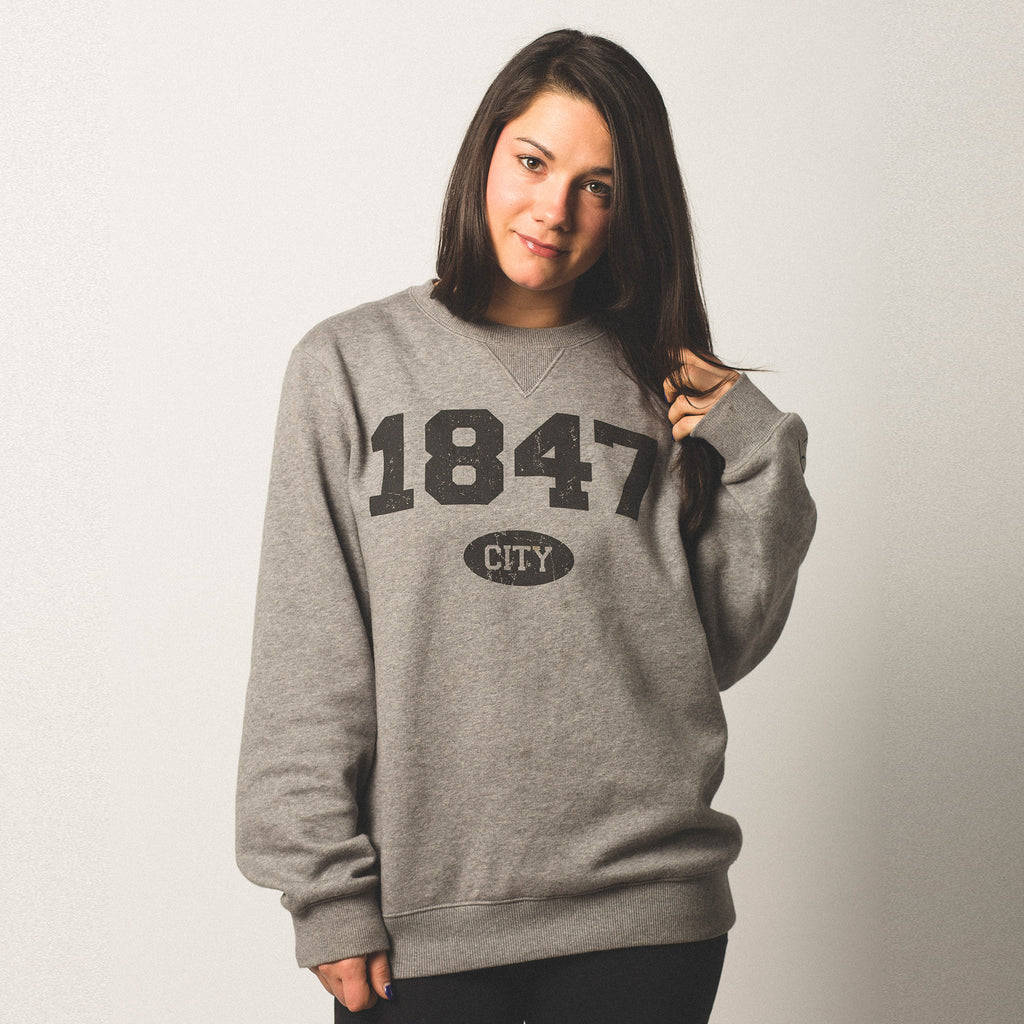 female model with olive skin and black hair wearing a vintage grey crewneck sweatshirt 1847 screen printed on front in black bold lettering city in small bubble underneath size small