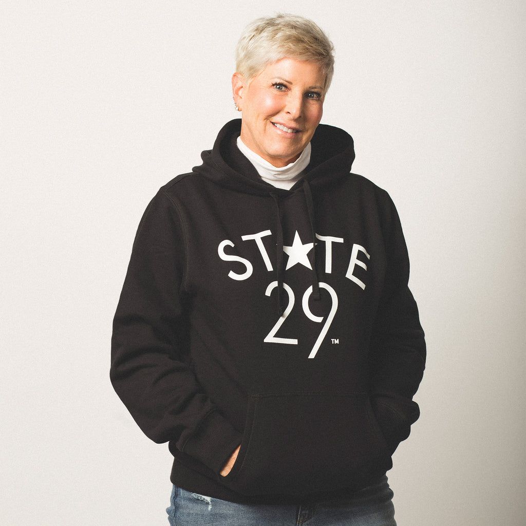 female model short blonde hair wearing a black pullover hooded sweatshirt featuring the 29th State Apparel logo trademarked in white on front above pouch