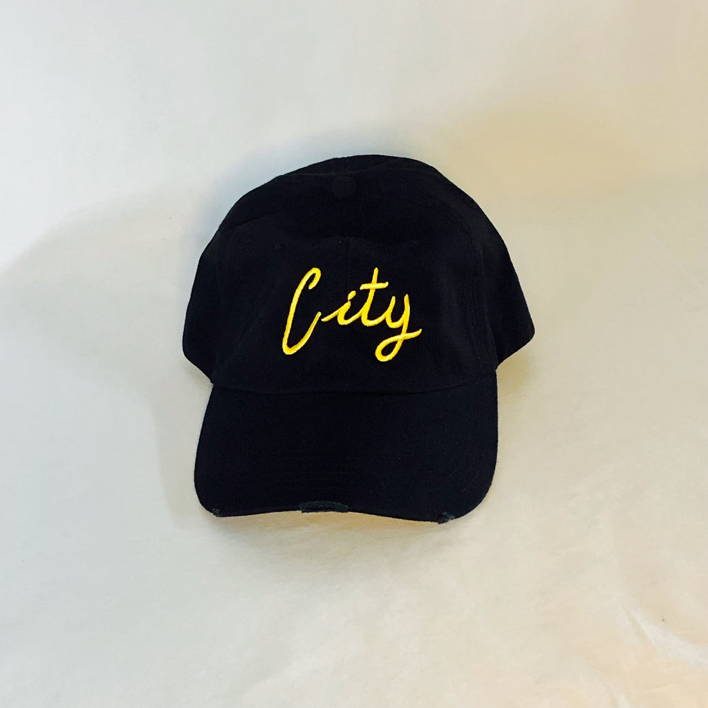all black 6-panel dad hat with stitched grommets and little bit of distressing on the front brim. "city" embroidered in fun, cursive font in gold threading