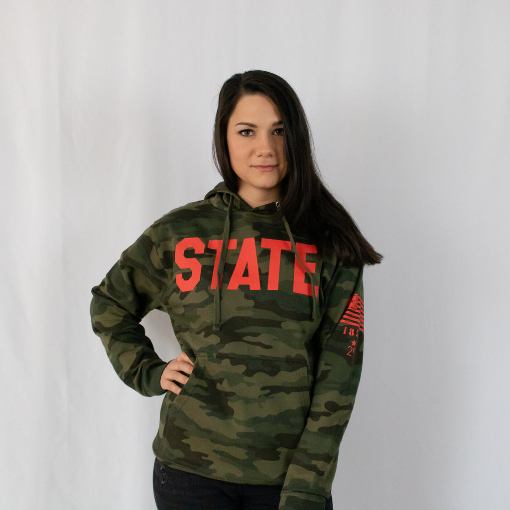 dark brown haired young woman in a military green camo hooded sweatshirt state printed in red on front red printed details on left sleeveright hand on hip