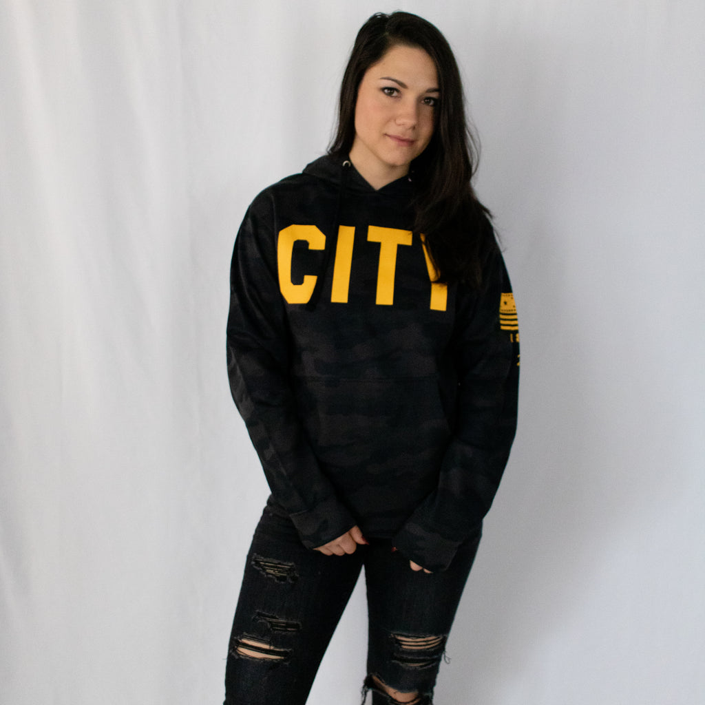 young woman with dark brown hair in black ripped jeans and a black military camo hoodie with city in gold printed across the chest