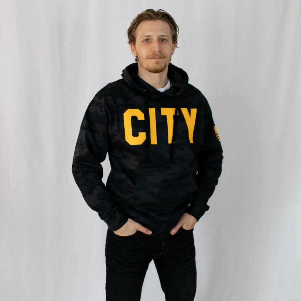 dirty blonde young man hands in his black jean pockets wearing a black military camo hoodie with city in gold printed on front center across chest